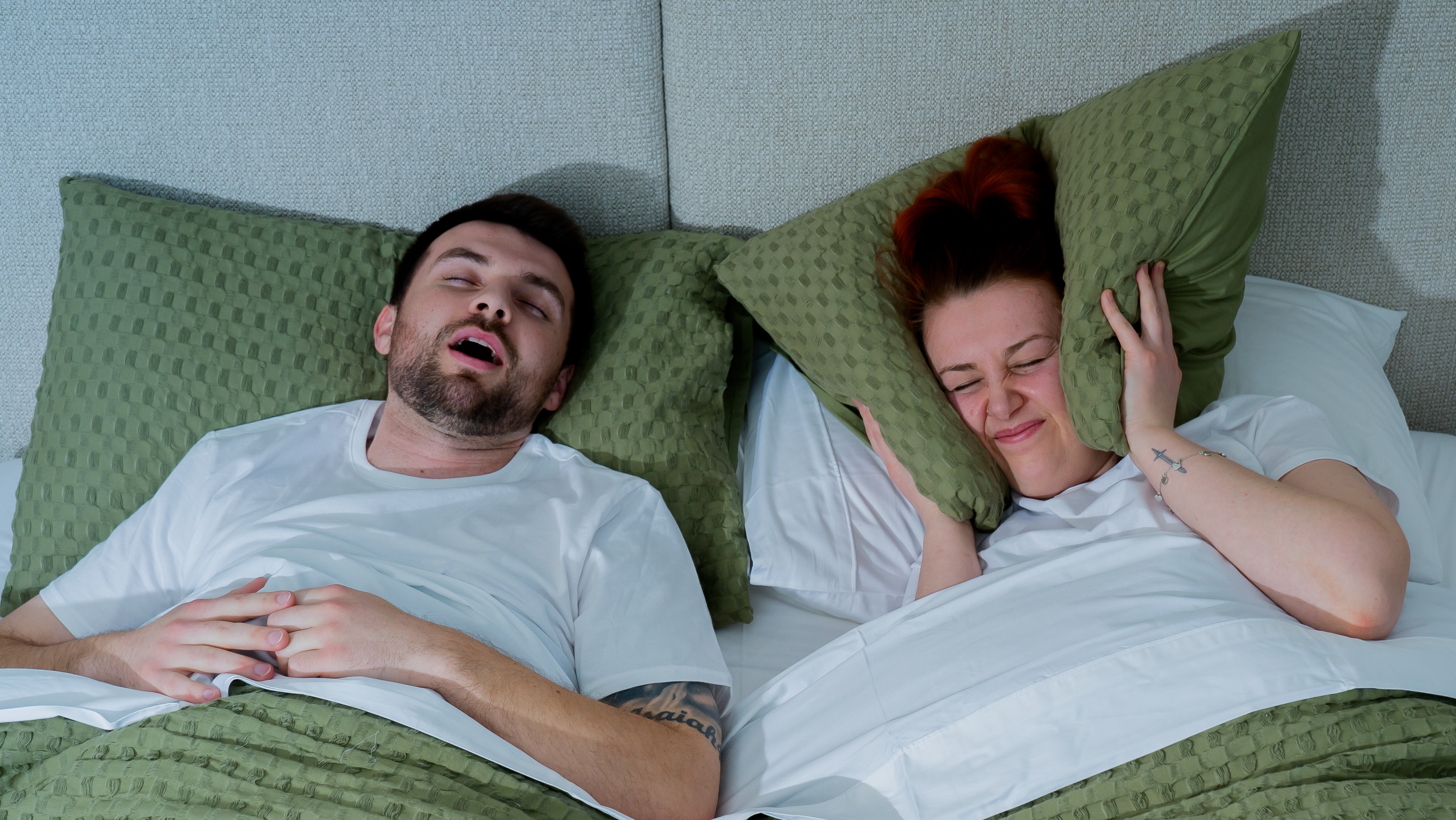 Man snoring loudly in bed while woman next to him looks frustrated and covers her ears with a pillow.